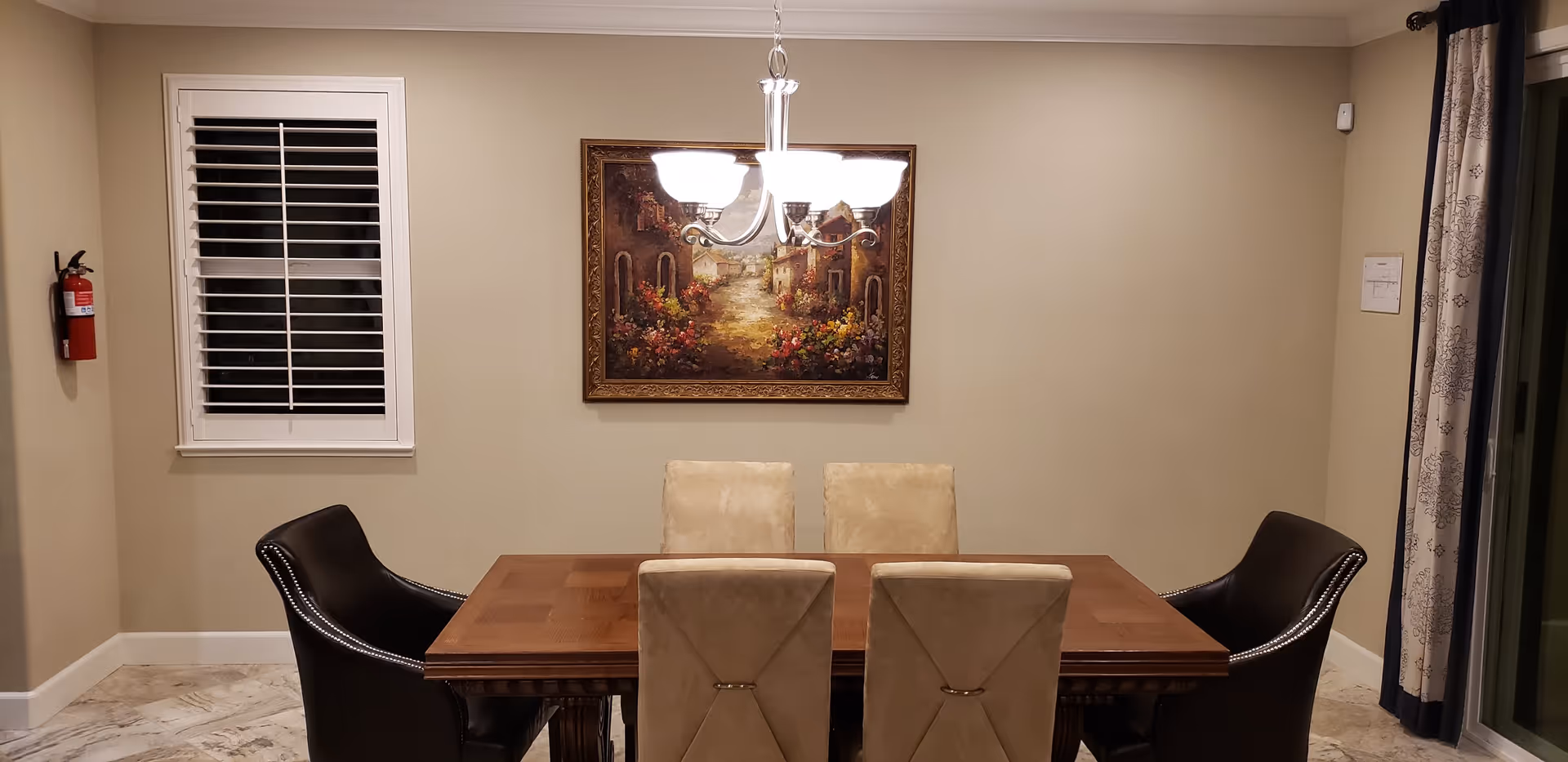 A dining room with a wooden table surrounded by six chairs, two black leather chairs on the ends and four beige cushioned chairs on the sides. A chandelier with five lights hangs above the table. On the wall behind the table is a framed painting depicting a scenic village street with flowers. To the left is a window with white shutters and a fire extinguisher mounted on the wall. To the right, there is a sliding glass door with patterned curtains.
