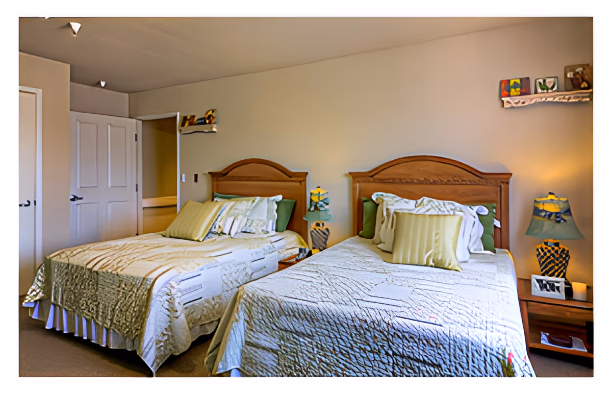 A bedroom with two twin beds featuring wooden headboards, cream-colored bedspreads, and multiple pillows. Between the beds is a small wooden nightstand with a decorative lamp and a framed photo. The room has beige walls, a door to the left, and two small shelves with decorative items mounted on the wall above the beds.
