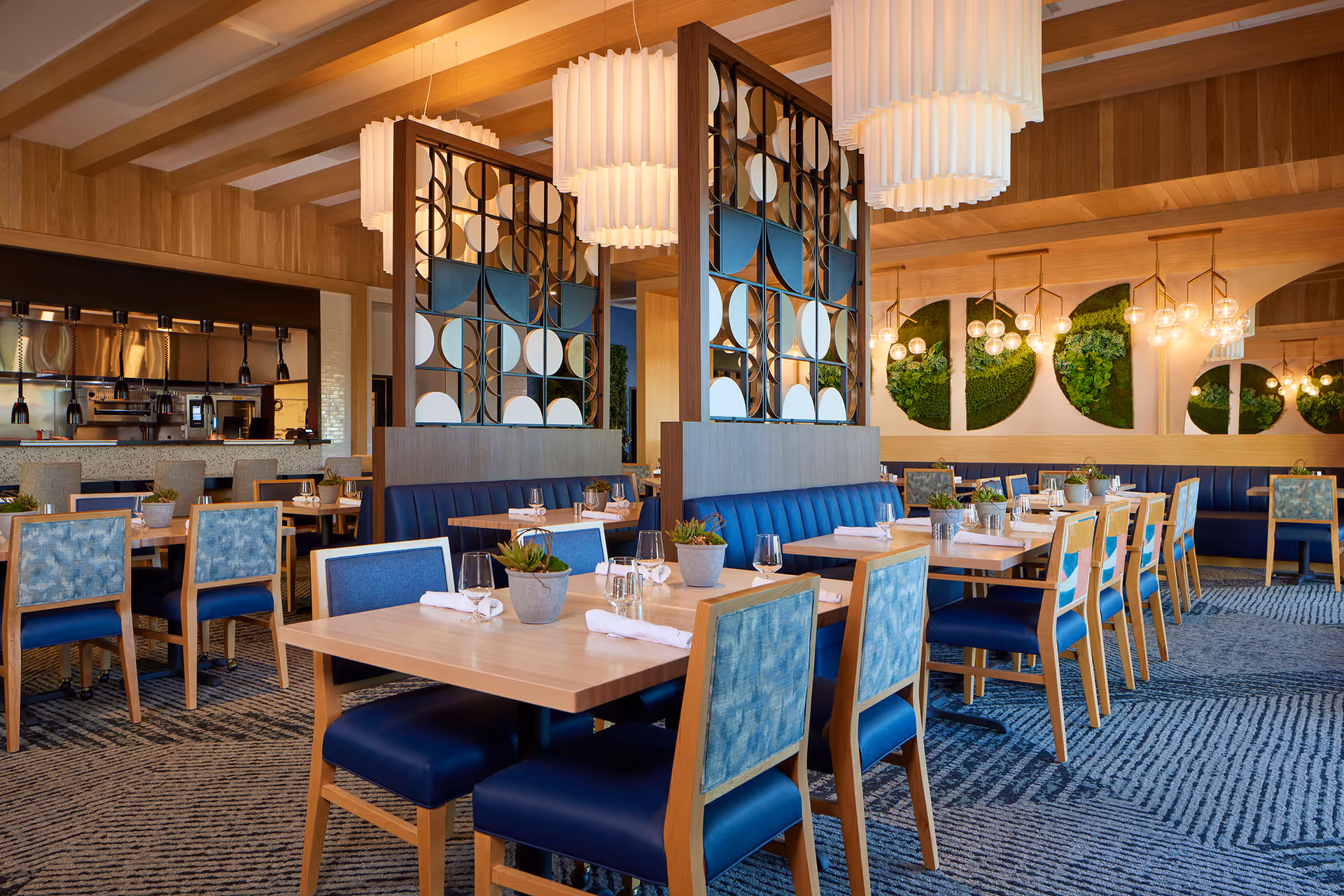 A modern dining room with wooden tables and blue cushioned chairs. The room features decorative partitions with circular patterns, hanging white pendant lights, and green wall art. The open kitchen area is visible in the background with bar stools along the counter.