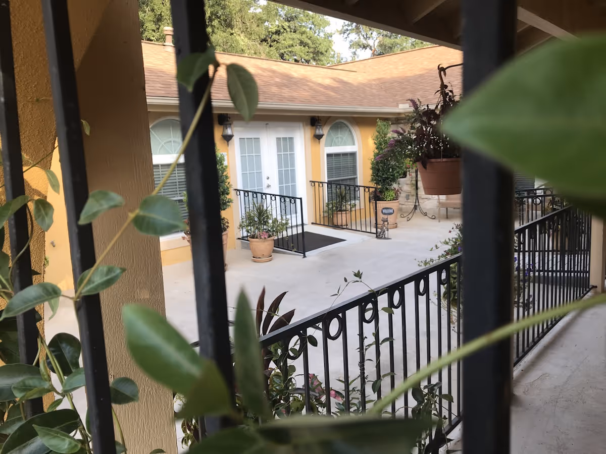 View through black metal bars of a courtyard area with a yellow building featuring white double doors and arched windows. The courtyard has potted plants, a hanging plant, and a black metal railing along a ramp leading to the doors. Trees are visible in the background.