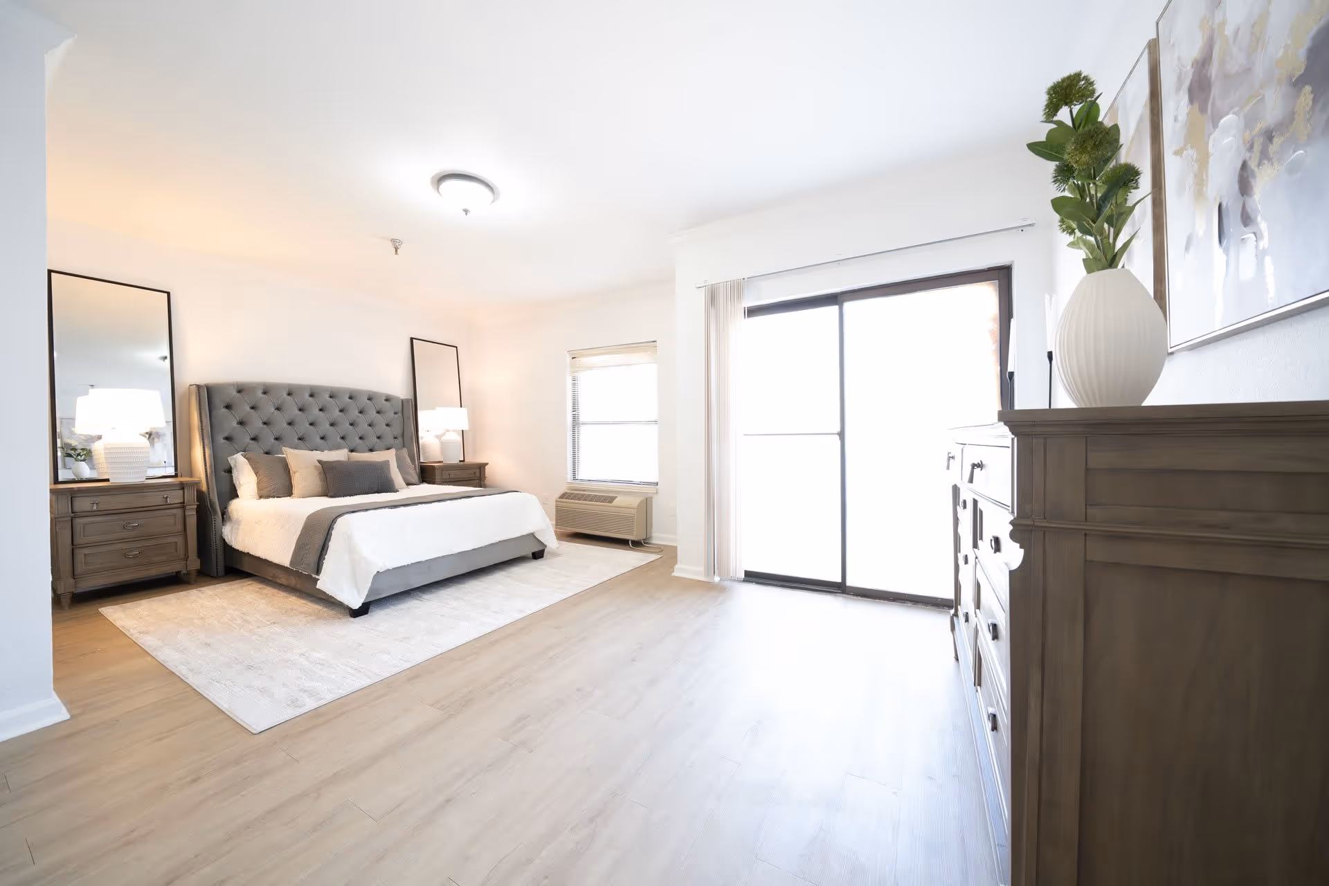 Bright, spacious bedroom with a tufted gray headboard, nightstands and lamps, a dresser, sliding glass door, and light wood flooring.