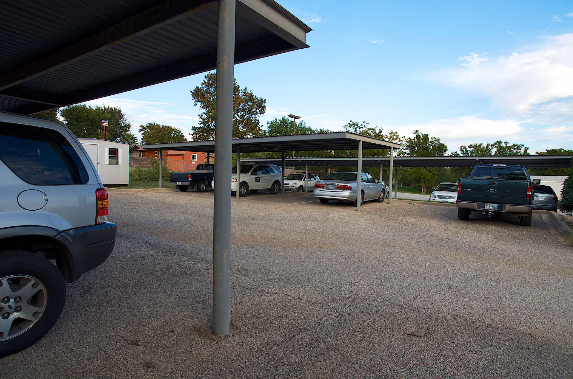 Covered parking area with several cars parked under metal carports in front of a building.