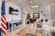 A bright and clean living room area with a white sofa, patterned armchair, glass coffee table, and a TV mounted on the wall. An American flag stands in the corner, and there is a large framed picture on the far wall. The room has tiled floors and a drop ceiling with fluorescent lighting.