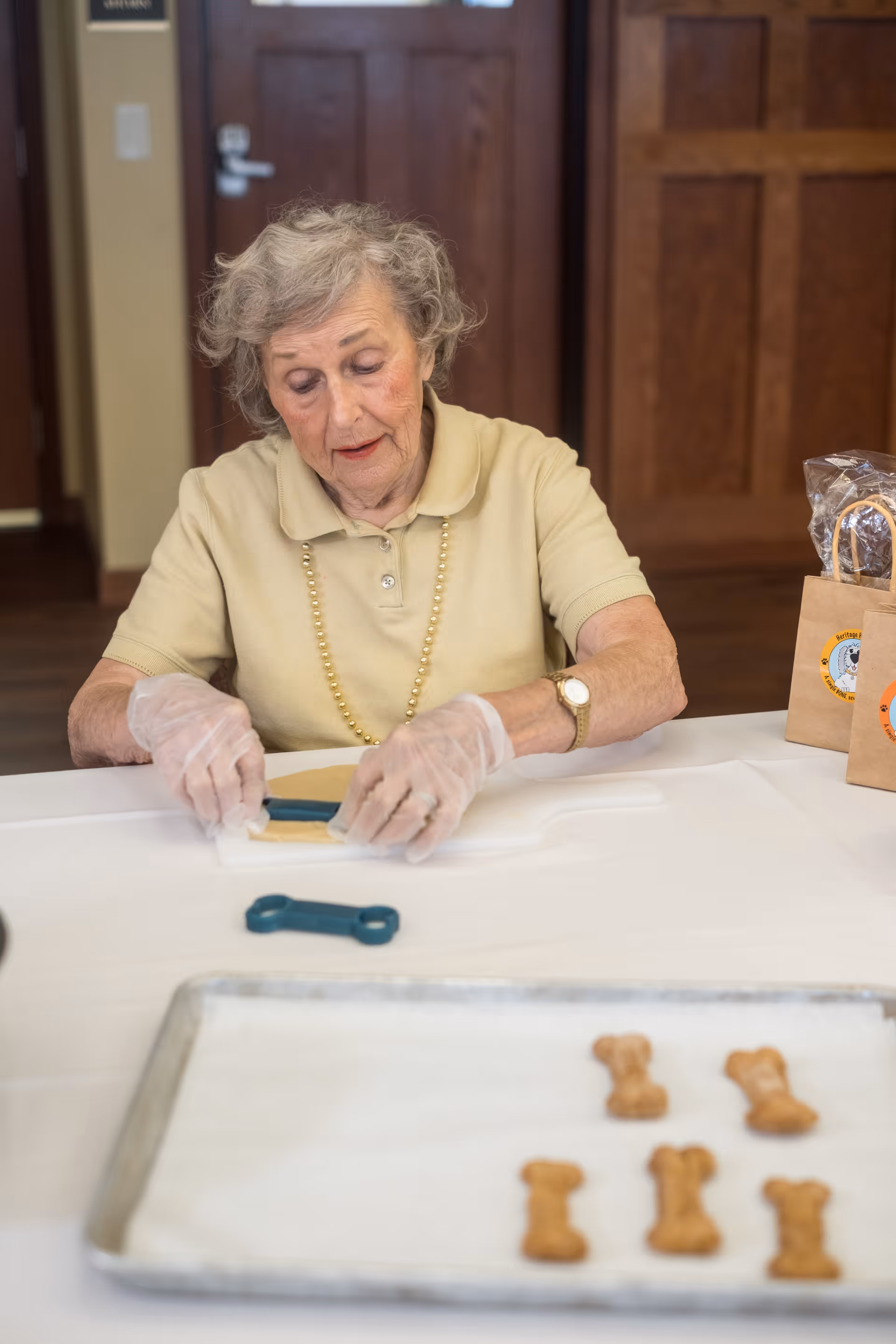 An elderly woman wearing a beige polo shirt and a pearl necklace is sitting at a table, using a blue bone-shaped cookie cutter on dough. She is wearing clear disposable gloves. In front of her on the table is a baking tray with several bone-shaped cookies. There are brown paper bags with a round logo on the right side of the table. The background shows wooden doors and a light switch.