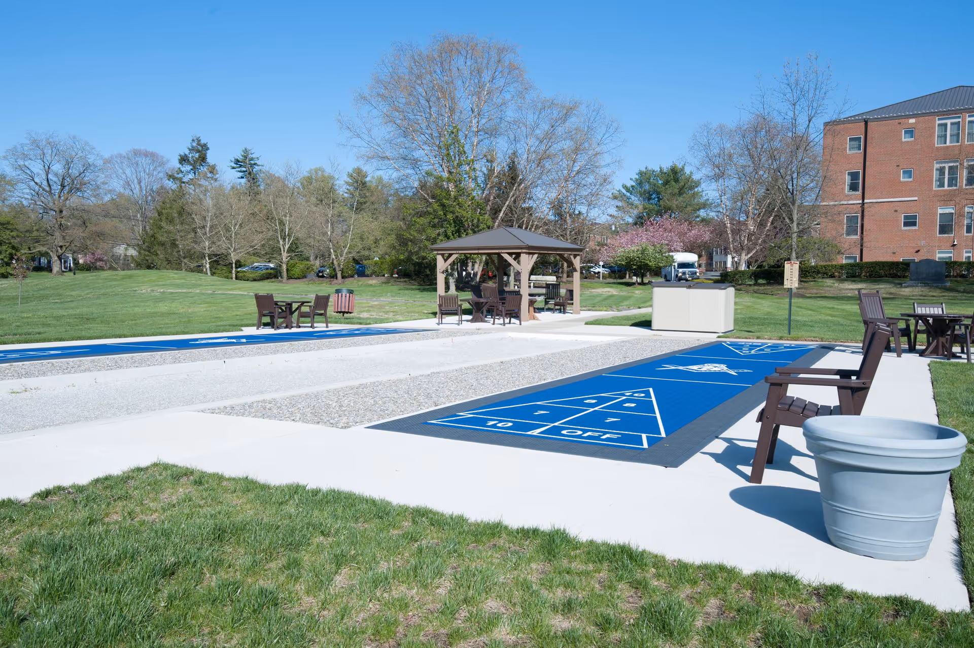 Outdoor shuffleboard courts with blue playing surfaces, surrounded by green grass and trees. There are wooden chairs and tables nearby, along with a small gazebo and a large brick building in the background under a clear blue sky.