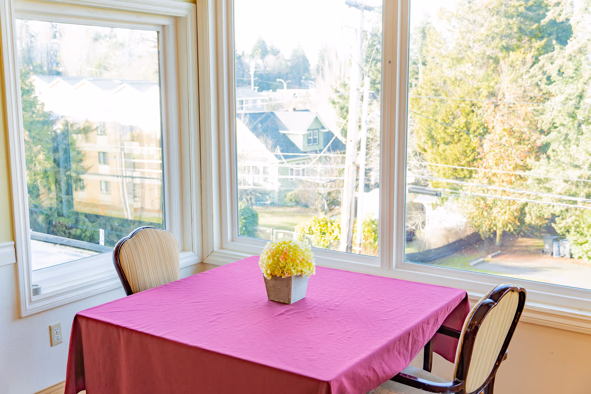A small dining area with a square table covered with a pink tablecloth and a small flower arrangement in the center. Two chairs with striped upholstery are placed on opposite sides of the table. Large windows surround the area, providing a view of trees and houses outside.