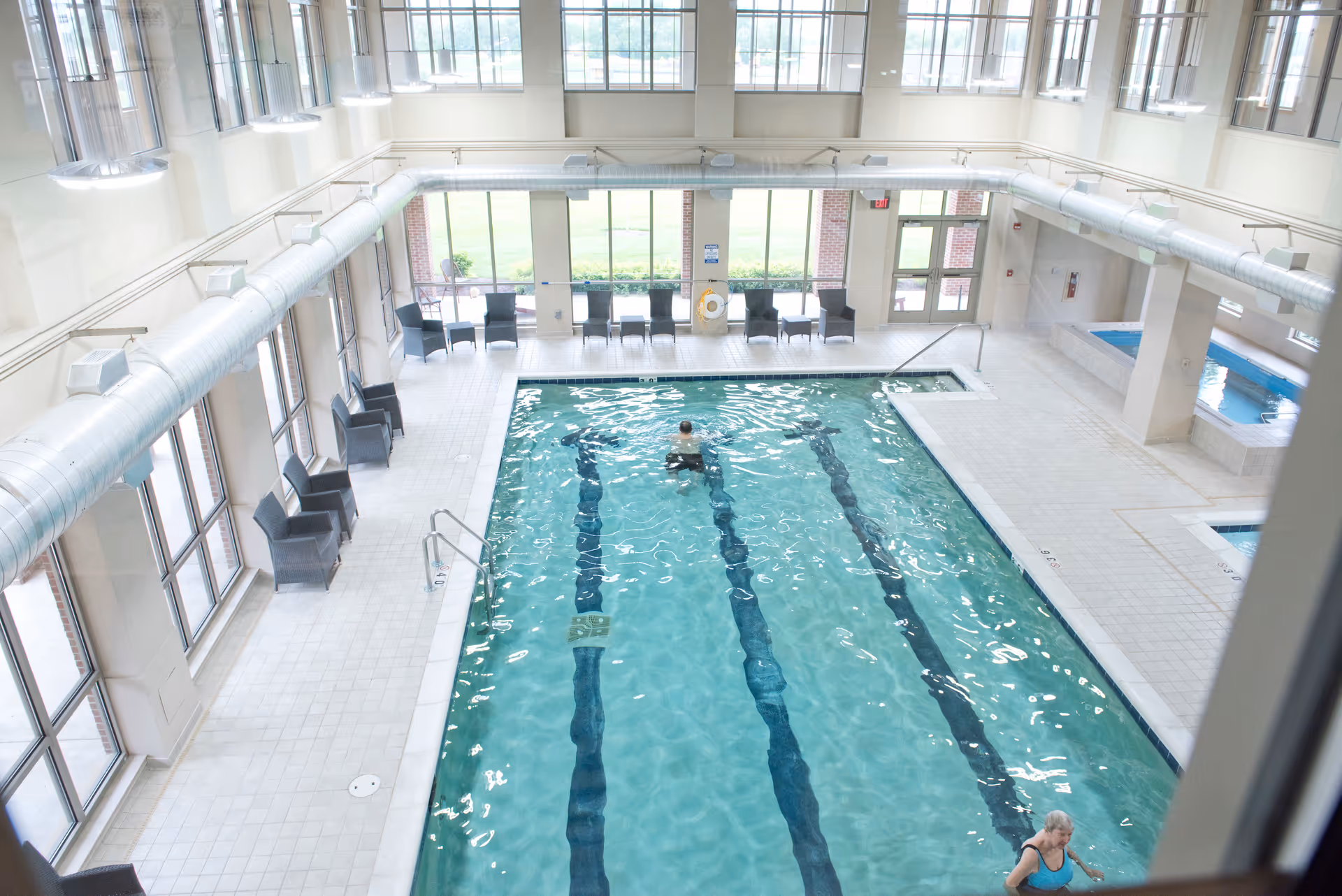 Indoor swimming pool with two people in the water, surrounded by large windows and several black chairs along the tiled pool deck. There is also a hot tub area visible to the right.
