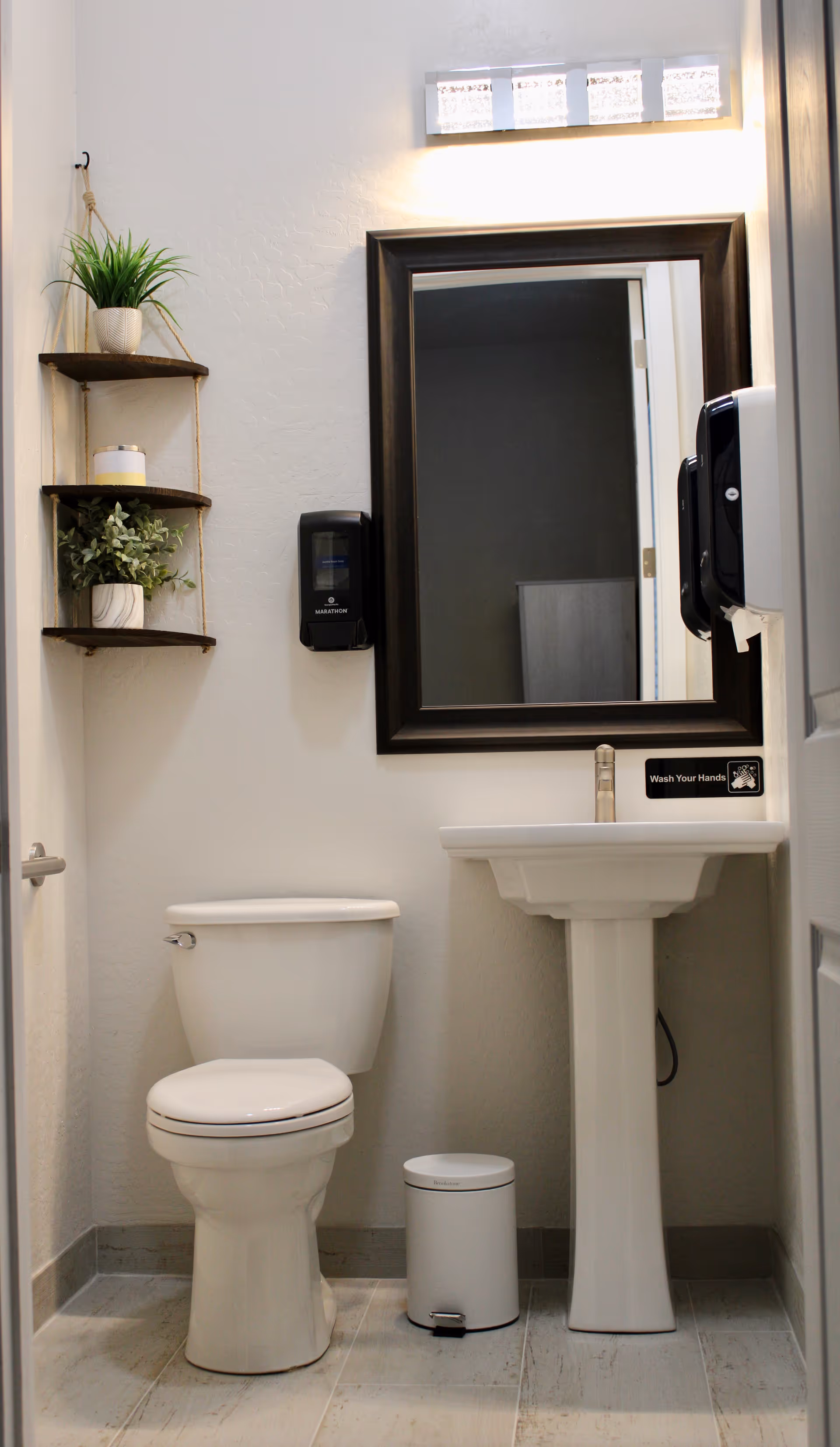 A clean and modern bathroom with a white toilet, a white pedestal sink, a large rectangular mirror with a dark frame, a small white trash can, and two wall-mounted soap dispensers. There is a small hanging shelf with two potted plants and a candle on the left wall.