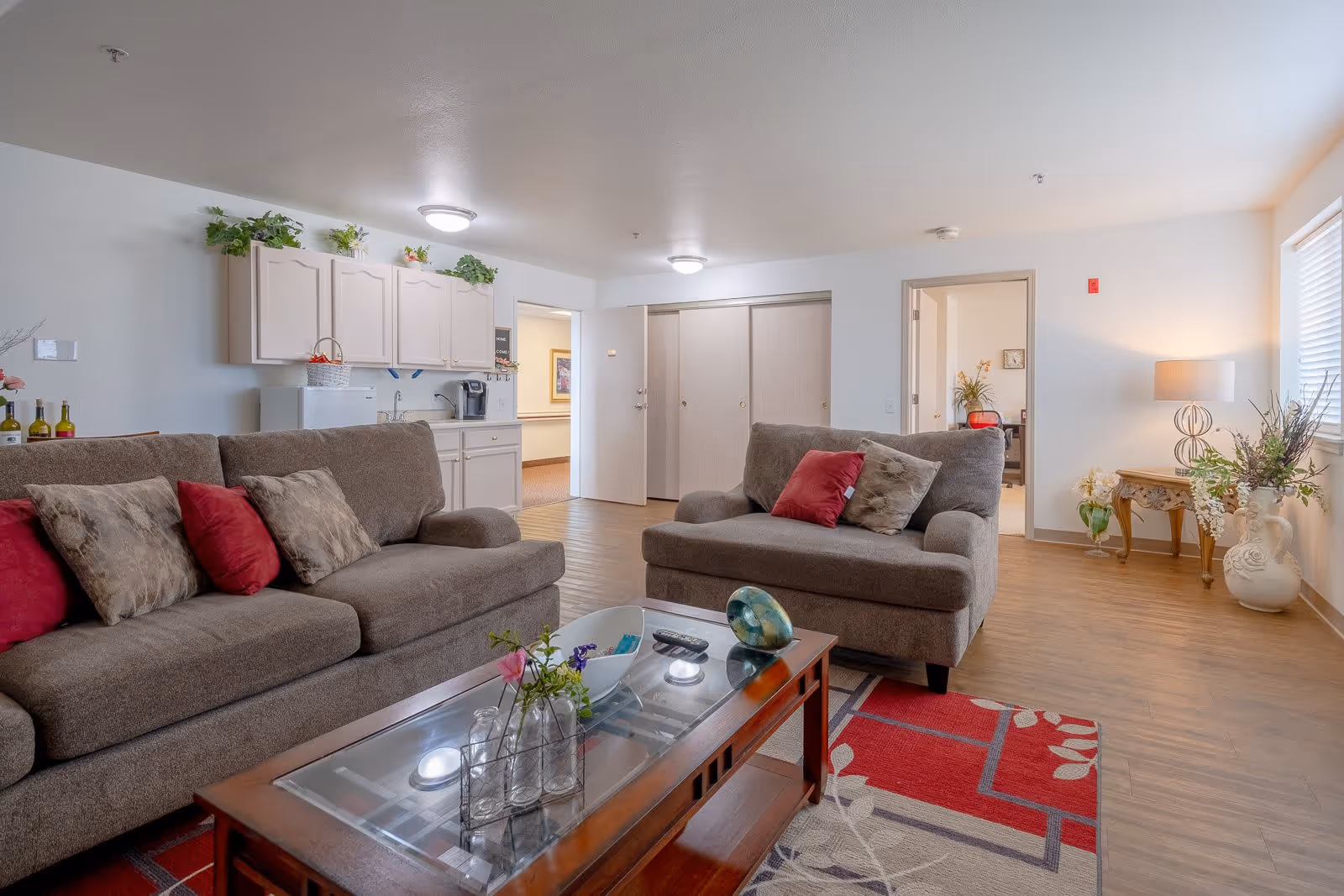 A cozy living room area with two brown sofas adorned with red and patterned pillows. A wooden coffee table with glass top holds decorative items and flowers. The room features light-colored walls, wood flooring, a small kitchenette with cabinets, a mini fridge, and a coffee maker. There is a doorway leading to another room and a window letting in natural light. Decorative plants and a table lamp add warmth to the space.