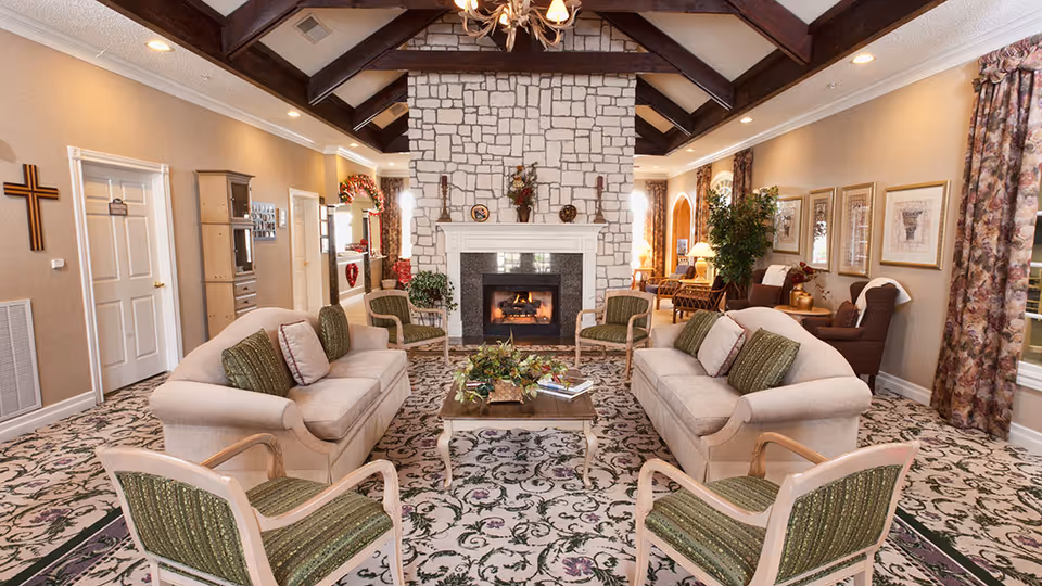 A cozy communal lounge with matching sofas and chairs arranged around a central coffee table and a stone fireplace under exposed wooden beams.
