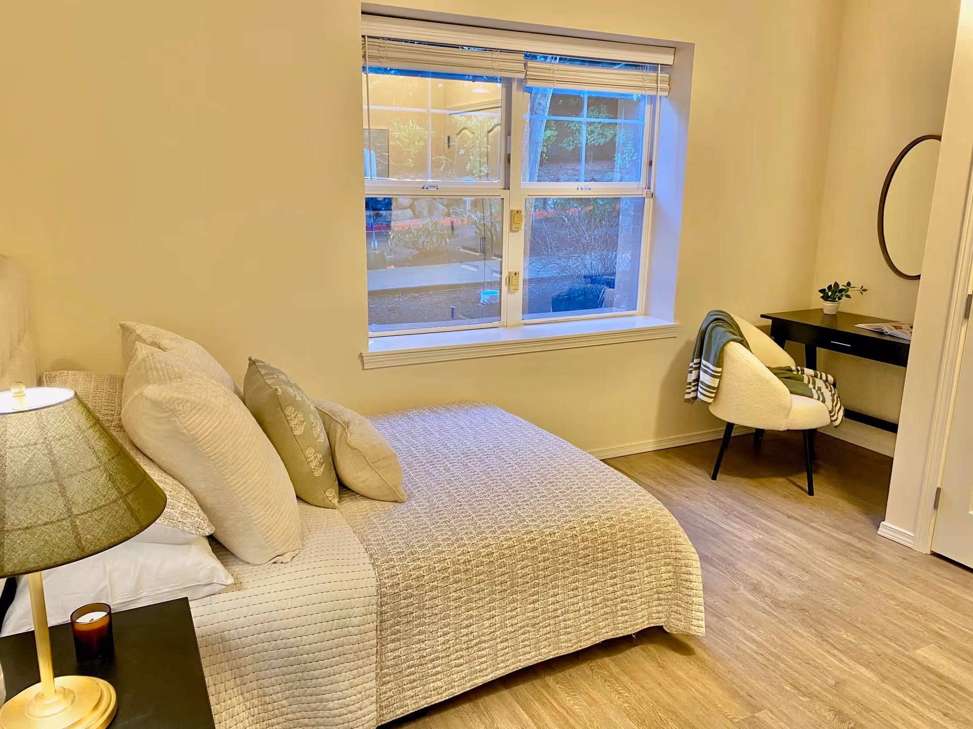 A cozy bedroom with a neatly made bed featuring multiple pillows and a textured beige bedspread. Next to the bed is a nightstand with a lamp and a small candle. A large window with white blinds lets in natural light, and a small desk with a round mirror and a cushioned chair with a green and white striped blanket is positioned against the wall. The room has light-colored walls and wood flooring.