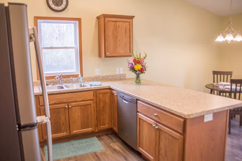 A kitchen with wooden cabinets, a double sink under a window, a stainless steel refrigerator, a dishwasher, and a countertop with a vase of colorful flowers. In the background, a dining area with a round table and chairs is visible under a hanging light fixture.