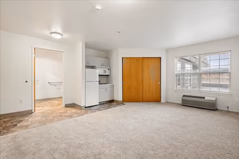 An empty apartment interior featuring a small kitchenette with white cabinets, a refrigerator, microwave, and sink. The room has beige carpet flooring, a large window with blinds, a wall-mounted air conditioning unit below the window, and a wooden double-door closet. The adjacent room has tiled flooring and a grab bar visible on the wall.