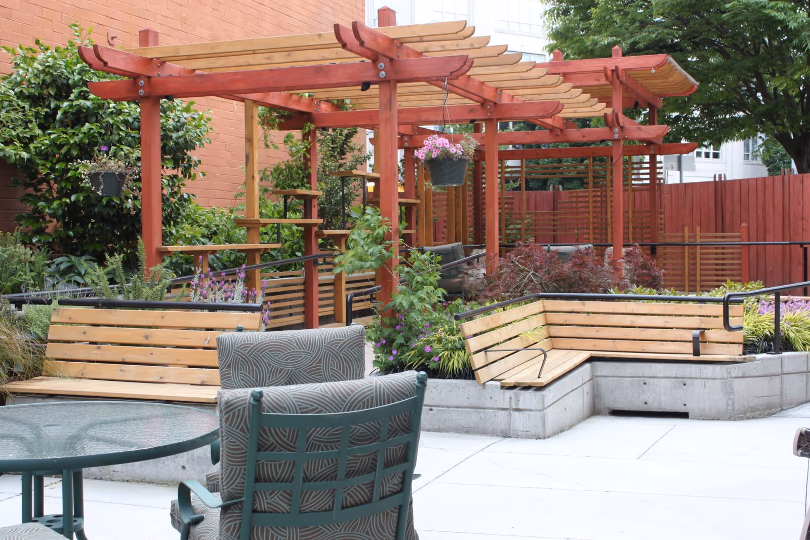 Outdoor courtyard with wooden pergola, planted garden beds, and wooden benches around a paved seating area.