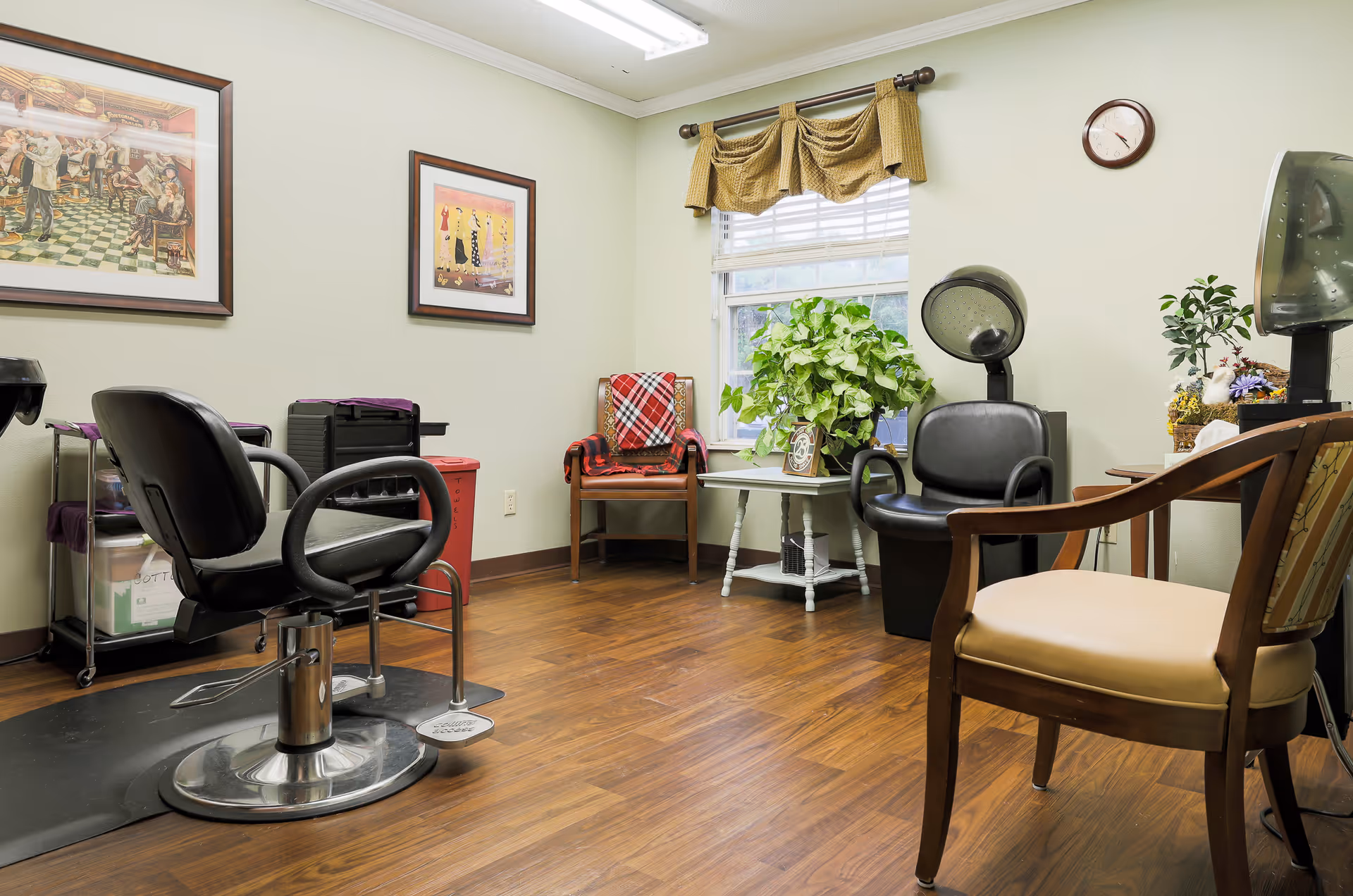 Interior of a small salon room with a black salon chair on a mat, a hair dryer chair, a wooden chair with a cushioned seat, a small table with a green leafy plant, and two framed pictures on the wall. The room has wooden flooring, a window with a valance, and a clock on the wall.
