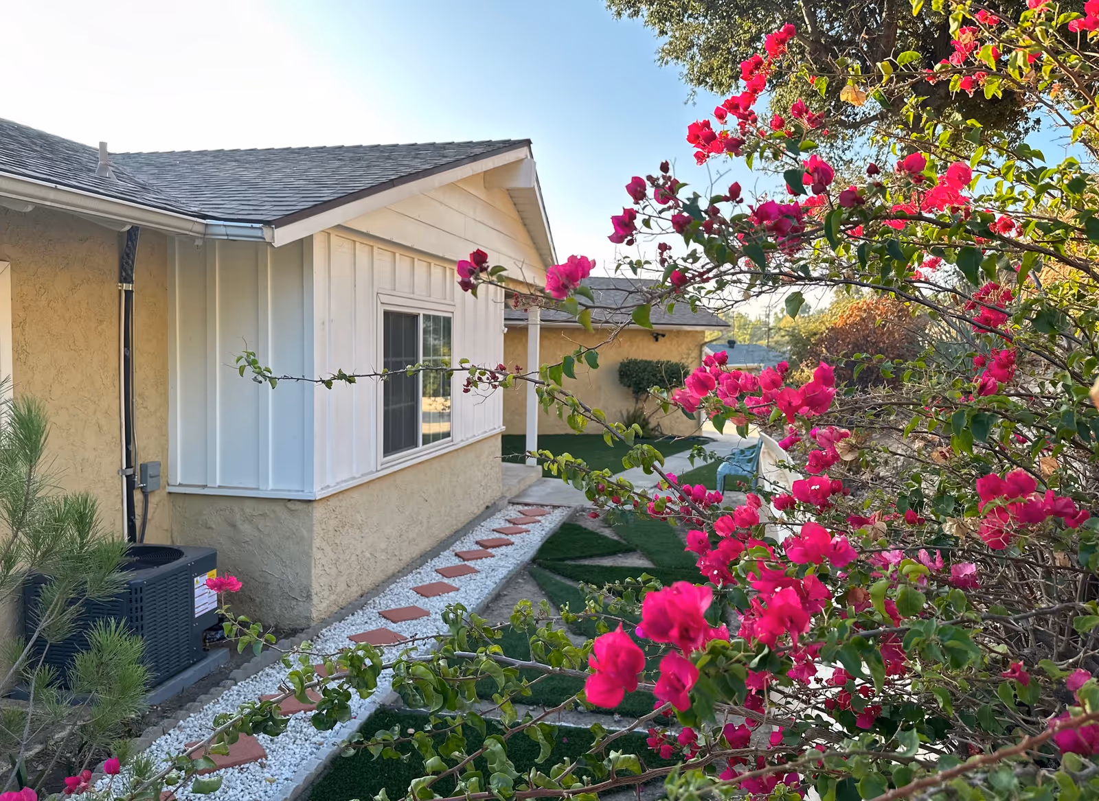 Well kept beige house exterior with white accents, stone walkway, and vibrant pink flowers in foreground.