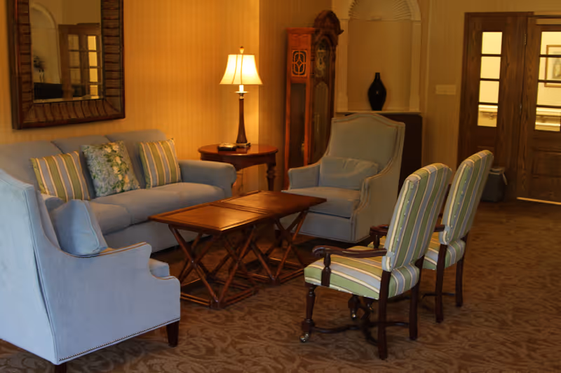 A cozy living room area with a light blue sofa, two matching armchairs, and two striped chairs arranged around a wooden coffee table. A table lamp on a side table and a tall grandfather clock are visible in the background, along with a large mirror on the wall and double wooden doors.