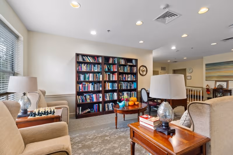 A cozy living room area in a senior living facility with beige armchairs, wooden side tables with lamps, a round coffee table with decorative pumpkins and flowers, and a large bookshelf filled with books against the wall. The room has a carpeted floor, a window with blinds, and soft overhead lighting.