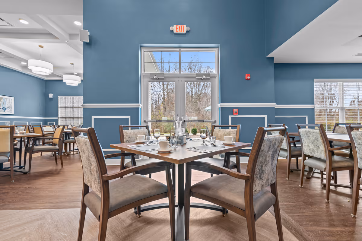 A bright and spacious dining room with blue walls and large windows letting in natural light. Several wooden tables are set with white cups, glasses, and silverware, surrounded by cushioned chairs. The room features modern light fixtures and a glass door leading outside, with a view of trees and greenery.