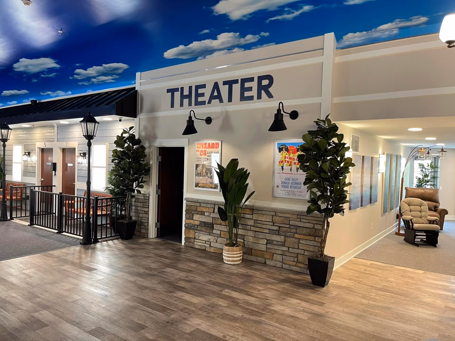Interior view of a memory care facility hallway with a theater entrance. The theater entrance has a sign above it that reads 'THEATER' and two movie posters on the wall. There are potted plants on either side of the entrance. To the left, there is a small fenced area with benches and doors numbered 5 and 7. The ceiling is painted with a blue sky and clouds. To the right, there is a hallway with a cushioned rocking chair and wall art.