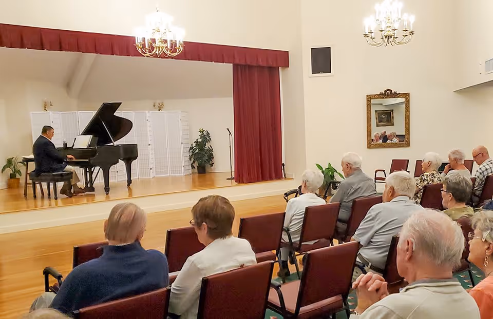 An elderly audience seated in chairs watching a man playing a grand piano on a stage with a red curtain backdrop in a well-lit room with chandeliers and a mirror on the wall.