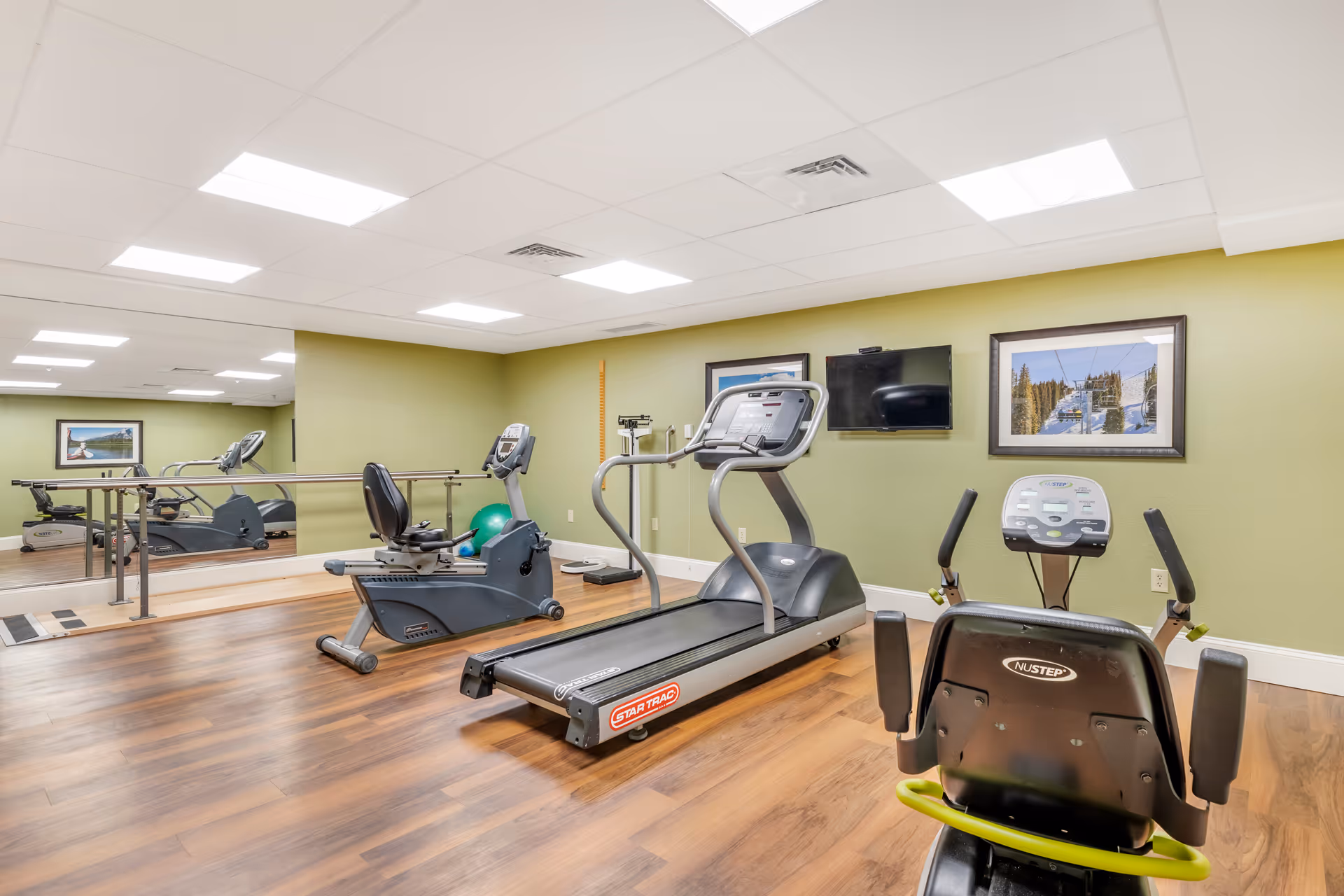 A small fitness room with wooden flooring and light green walls. The room contains exercise equipment including a treadmill, a recumbent bike, and another stationary bike. There is a large mirror on one wall, a flat-screen TV mounted on the wall, and two framed pictures depicting outdoor scenes. The ceiling has recessed lighting panels.