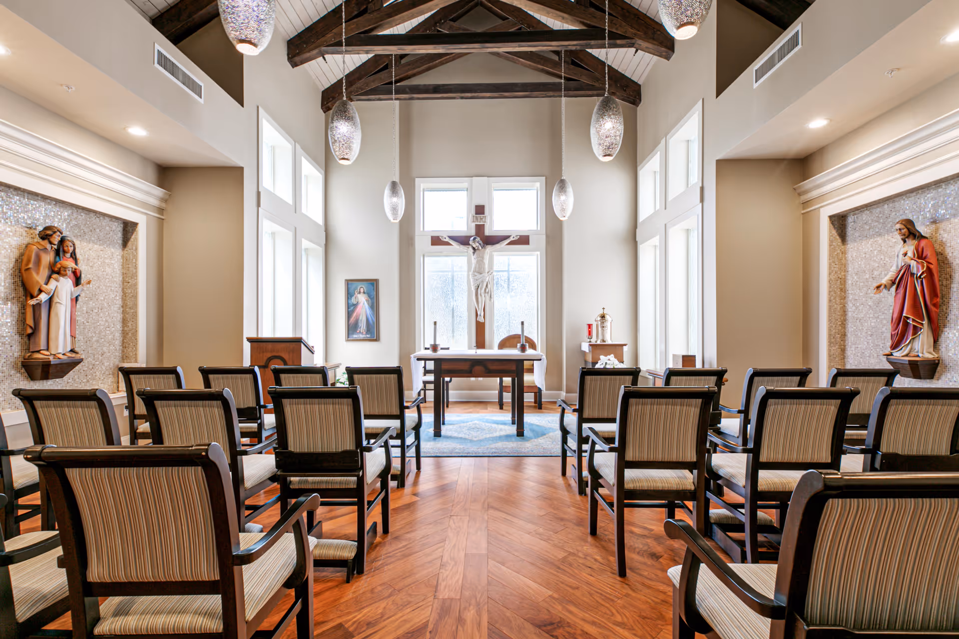 Interior view of a chapel with wooden beams on the ceiling, pendant lights, rows of chairs with striped upholstery facing an altar. The altar has a crucifix with Jesus, candles, and religious statues on either side set in mosaic niches.
