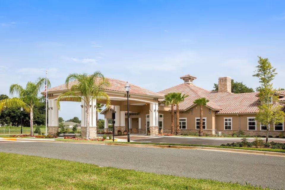 Exterior view of Mission Oaks Assisted Living and Memory Care facility showing a single-story building with a tiled roof, a covered entrance supported by columns, palm trees, and a well-maintained lawn and landscaping under a clear blue sky.