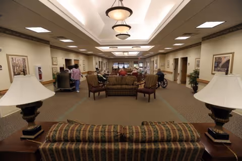 A spacious and well-lit common area in a senior living facility with multiple couches and chairs arranged for socializing. Several elderly residents and staff members are visible in the background. The room features beige walls, framed artwork, carpeted floors, and ceiling lights with decorative fixtures.