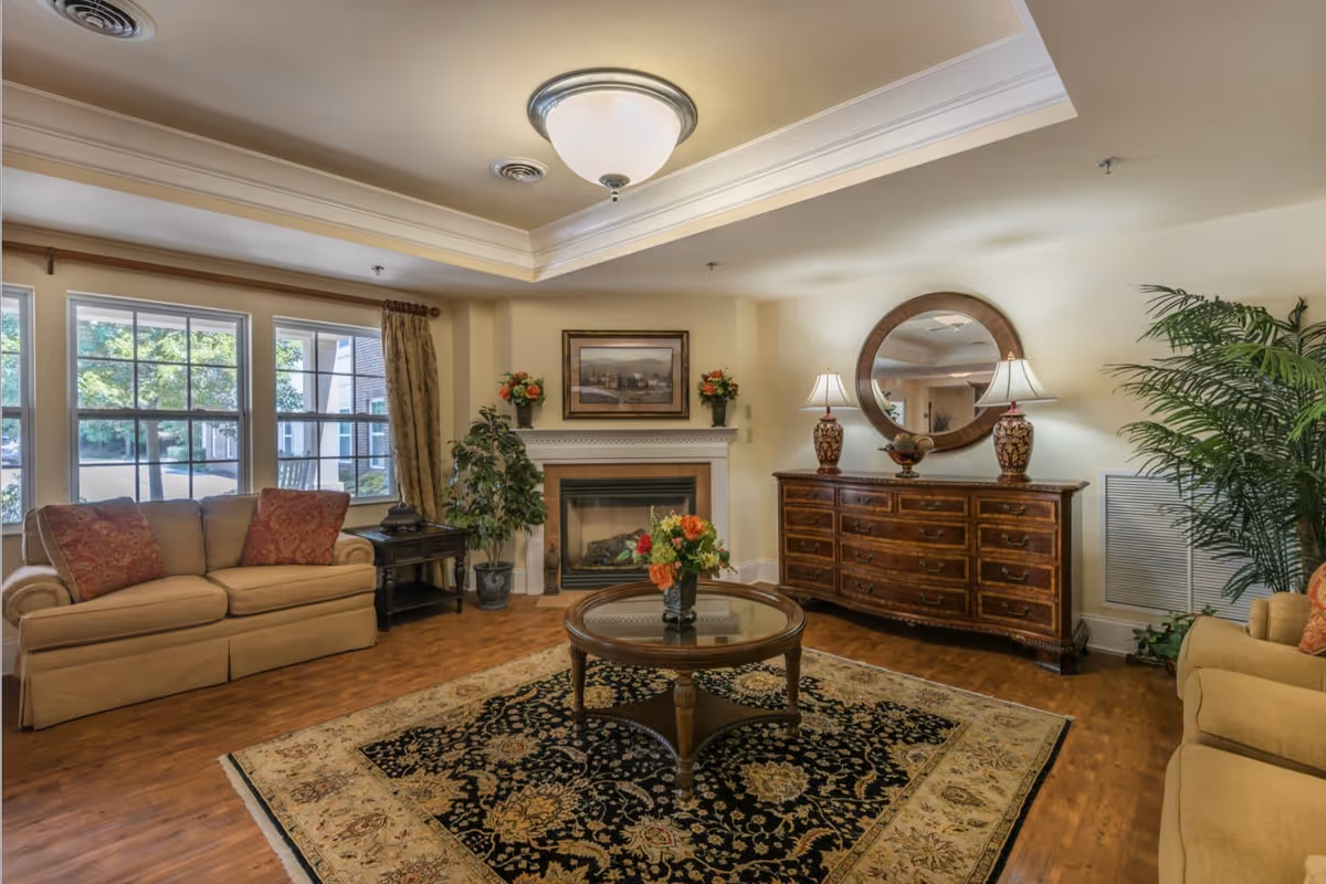 A cozy living room with two beige sofas adorned with red patterned cushions, a wooden coffee table with a glass top and a floral arrangement, a large ornate area rug, a wooden dresser with two decorative lamps and a round mirror above it, a fireplace with a framed painting and flower vases on the mantel, and large windows letting in natural light.