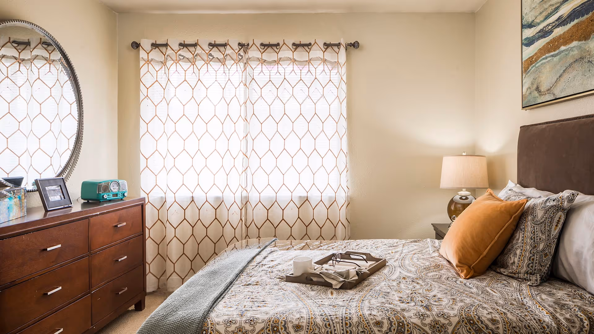 A cozy bedroom with a neatly made bed featuring patterned bedding and multiple pillows, including an orange one. A wooden dresser with a round mirror, a small framed photo, and a retro-style green radio sits to the left. Light filters through patterned curtains covering a window behind the bed. A bedside table with a lamp is on the right side of the bed, and a piece of abstract artwork hangs on the wall above the headboard.