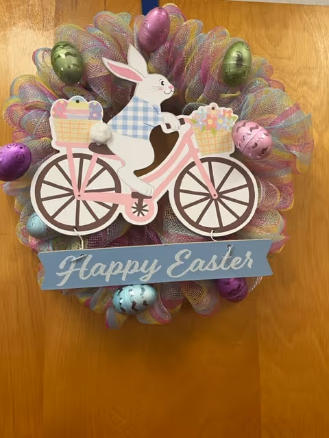 A colorful Easter wreath hanging on a wooden door. The wreath features a decorative white bunny wearing a blue checkered shirt riding a pink bicycle with baskets filled with Easter eggs and flowers. Surrounding the bunny are pastel-colored mesh ribbons and metallic Easter eggs in purple, green, and blue. A blue sign at the bottom of the wreath reads 'Happy Easter'.
