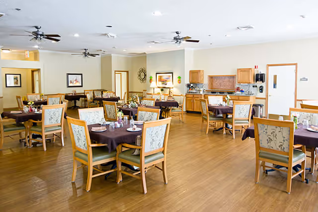 A spacious dining room with multiple wooden tables covered with dark tablecloths and set with plates, glasses, and silverware. The room has wooden flooring, ceiling fans, framed artwork on the walls, and a small kitchenette area with cabinets and a countertop in the background.