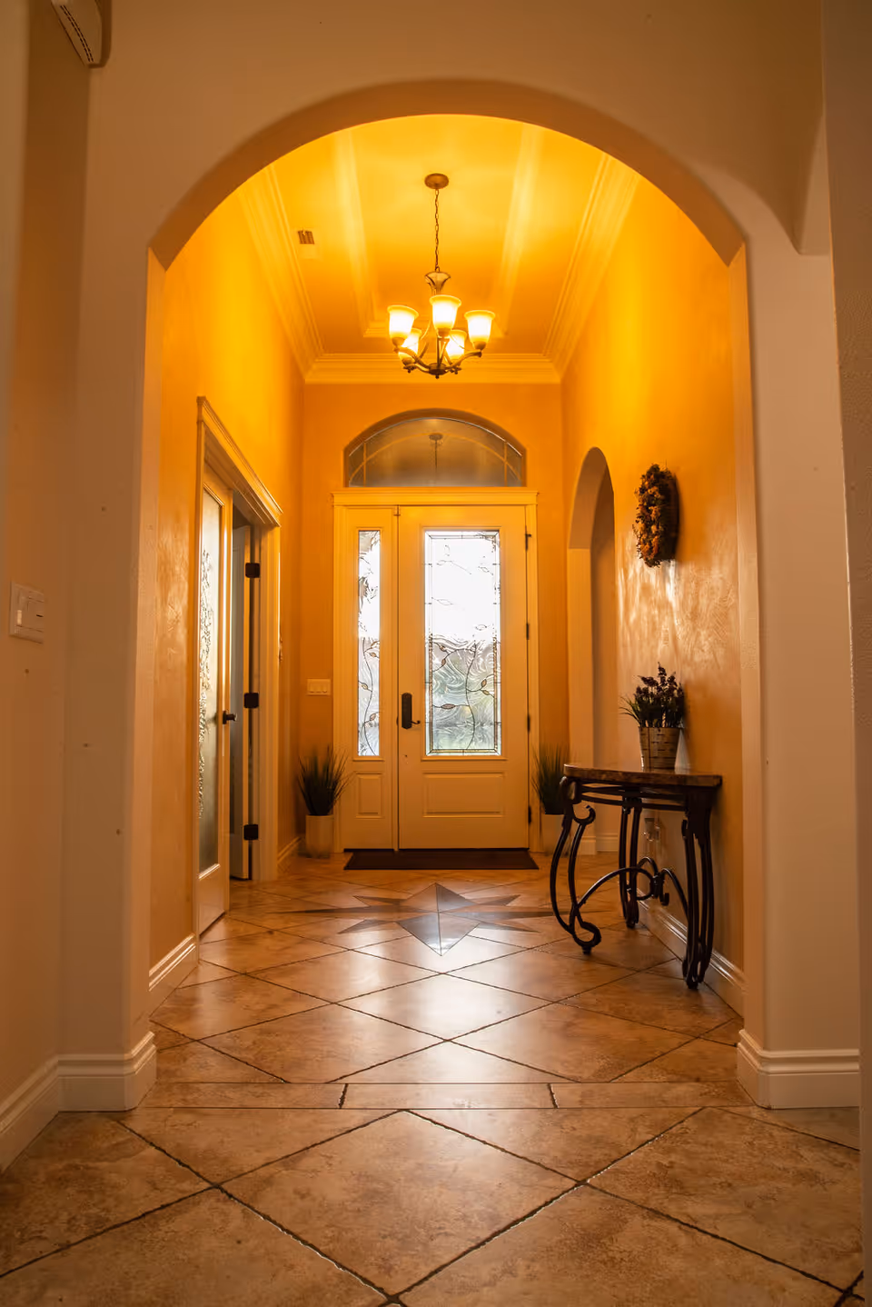 Warm-lit tiled entry hallway with an arched ceiling leading to decorative double front doors.