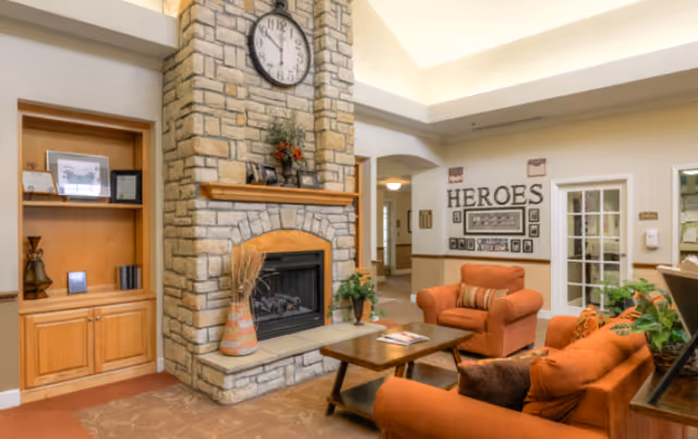A cozy living room area in Carroll Place featuring a stone fireplace with a clock above it, two orange upholstered armchairs, a wooden coffee table, built-in wooden shelves with decorative items, and a wall display with the word 'HEROES' and framed pictures.