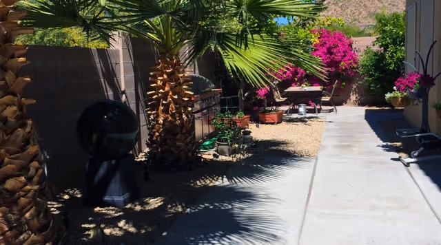 A sunny outdoor pathway lined with palm trees and various potted plants, including vibrant pink flowers. There is a barbecue grill along the left side near the wall and a small seating area with a table and chairs further down the path. The area is surrounded by a low wall and mountains are visible in the background.