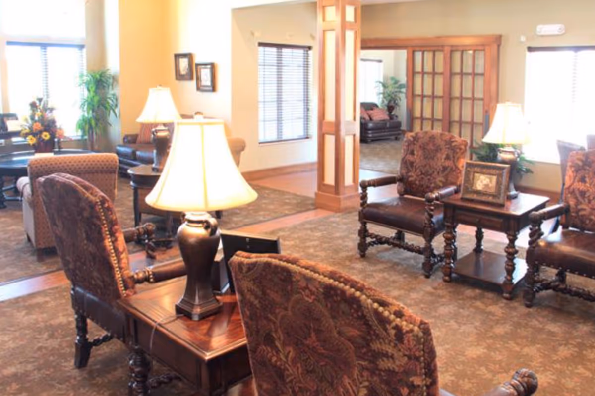 A well-lit living room area with patterned upholstered chairs arranged around wooden side tables with lamps. The room features large windows with blinds, framed pictures on the walls, and plants in the corners. There is a carpeted floor and wooden accents around the room, including a wooden column and French doors leading to another room.