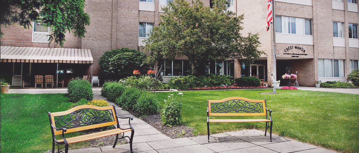 Outdoor courtyard area of Crest Manor Living & Rehabilitation Center featuring two decorative benches on a paved walkway, green grass, bushes, flowers, and trees. The building facade with windows and an entrance with a sign reading 'Crest Manor Living & Rehabilitation Center' is visible in the background along with an American flag.