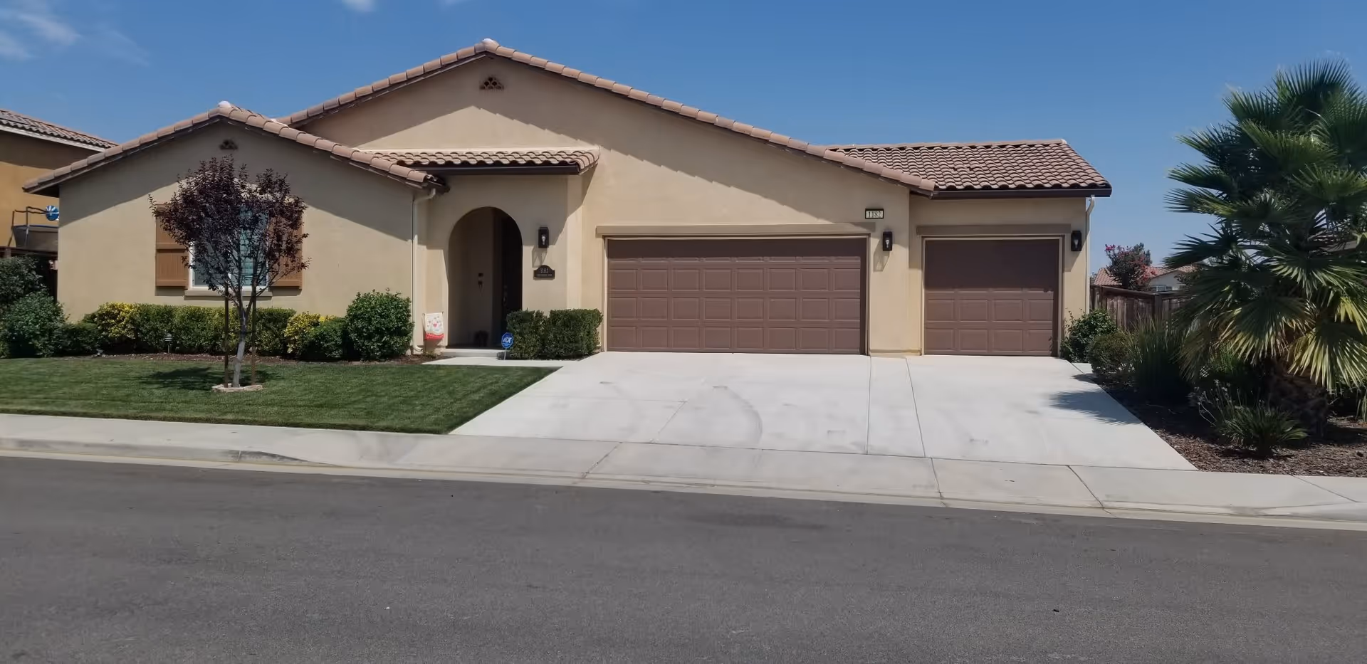 Front exterior view of a single-story house with beige stucco walls and a tiled roof. The house features a three-car garage with brown doors, a small front lawn with a tree and bushes, and a concrete driveway. The sky is clear and blue.