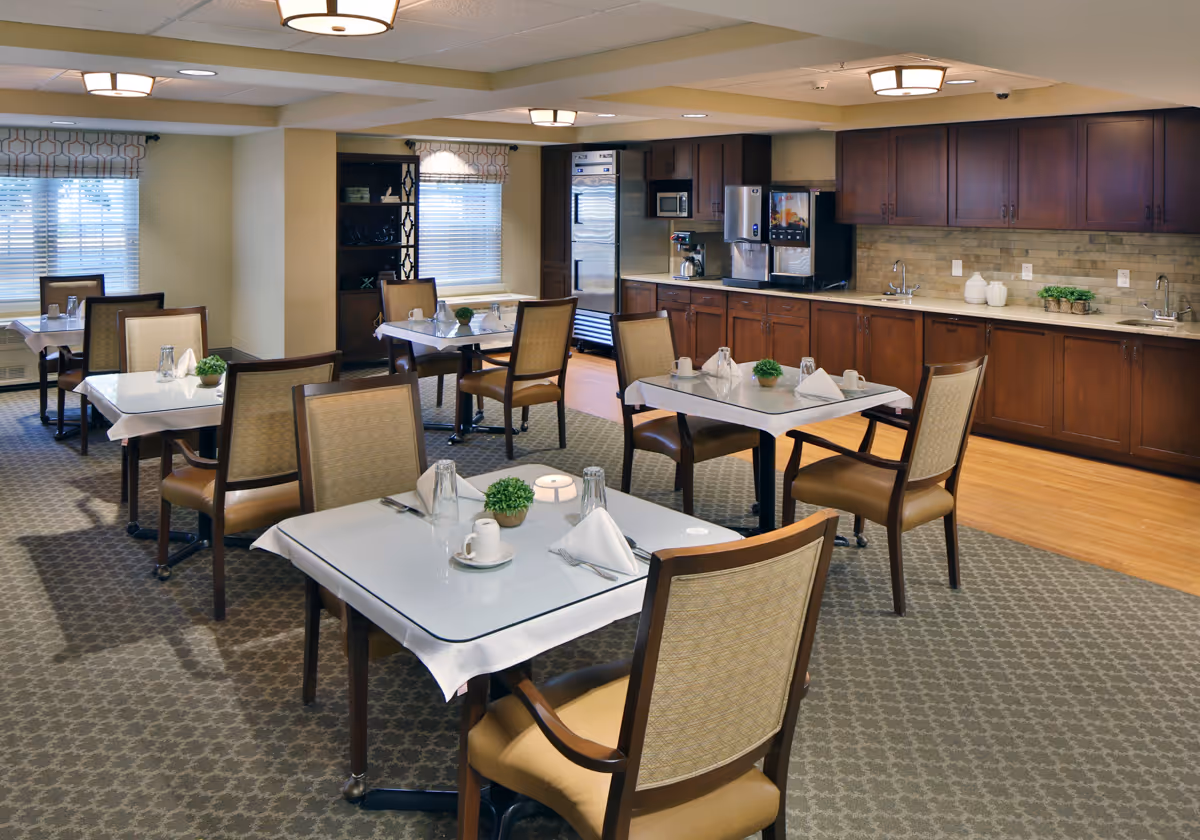 Well-lit dining room with multiple set tables and chairs and a kitchenette with cabinets along the back wall.