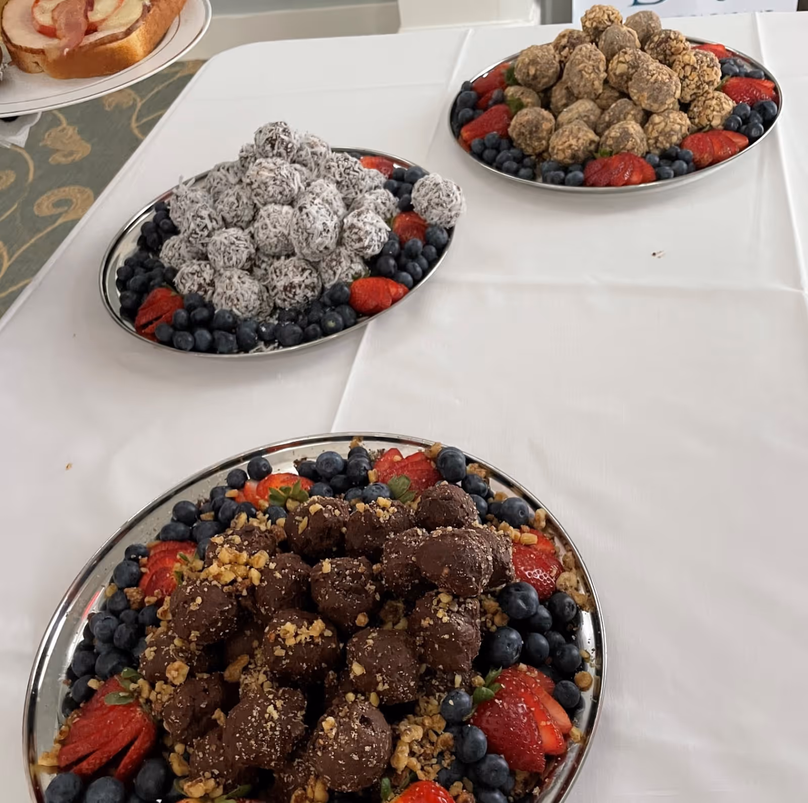 Three silver platters on a white tablecloth, each filled with different types of dessert balls surrounded by fresh blueberries and sliced strawberries. In the background, part of a sandwich on a plate is visible.