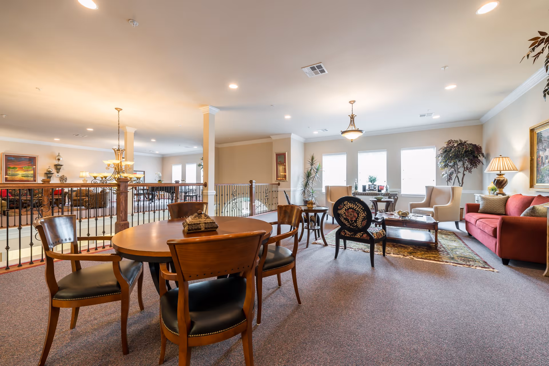 A spacious senior living common area with a round wooden table and four chairs in the foreground. The room features a seating area with a red sofa, two beige armchairs, a floral upholstered chair, a coffee table, and decorative plants. Large windows allow natural light to fill the room, and warm lighting fixtures hang from the ceiling. The space has a carpeted floor and neutral-colored walls with framed artwork.