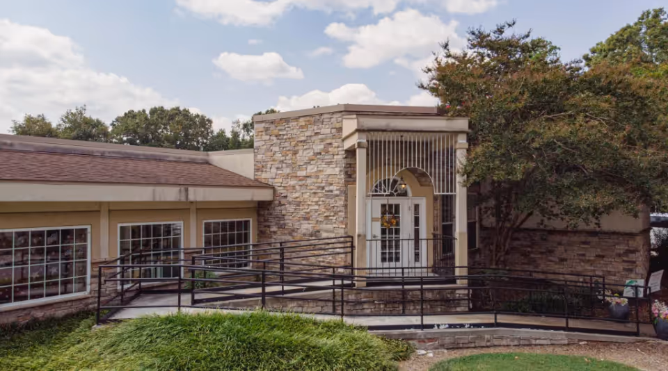 Stone-faced building entrance with double glass doors, a metal accessibility ramp, and surrounding landscaping.