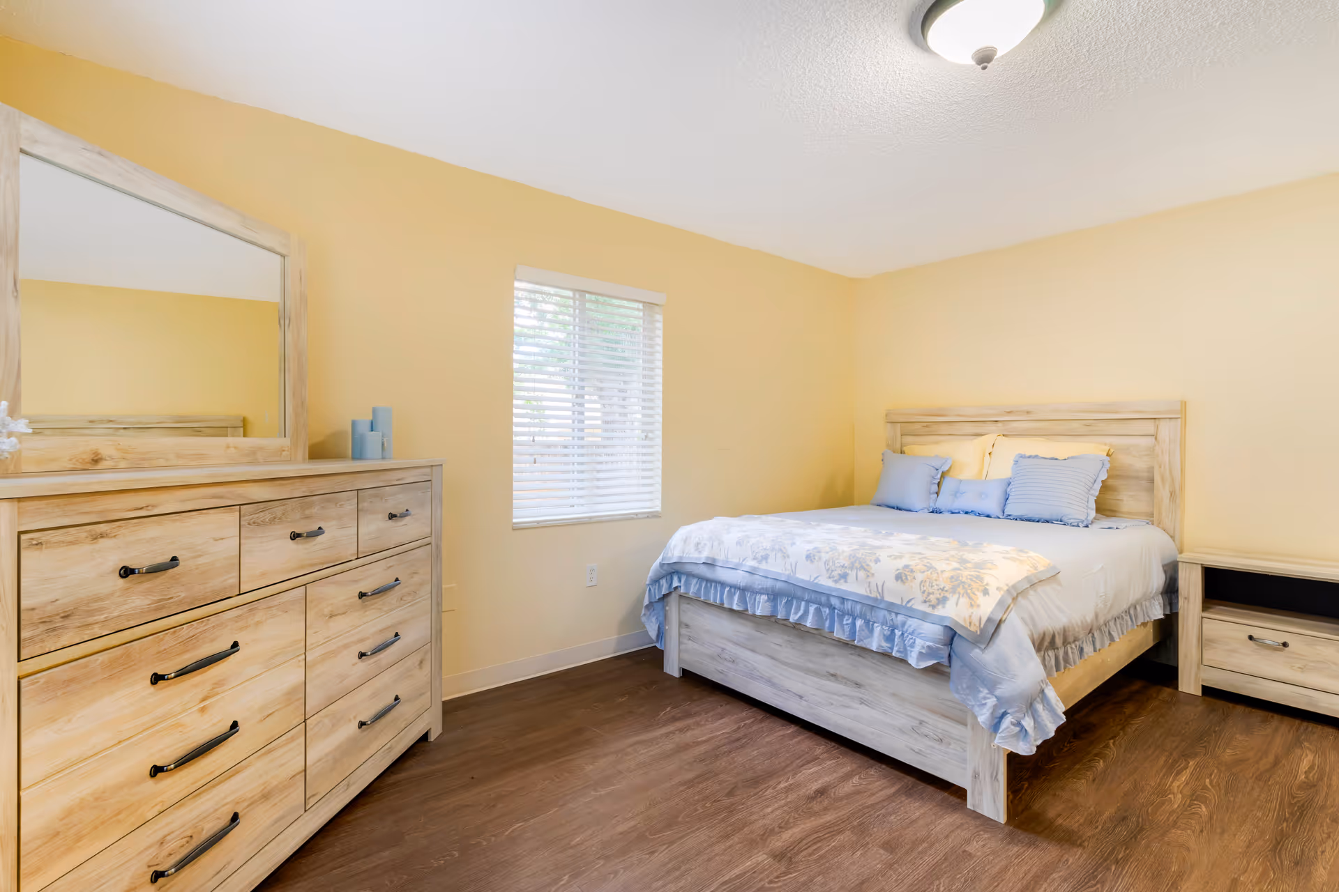 A bedroom with light yellow walls and wood flooring featuring a wooden bed with a floral bedspread and blue pillows. There is a matching wooden dresser with a large mirror and a nightstand beside the bed. A window with white blinds is on the wall behind the bed.