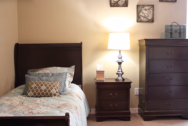 A bedroom with a dark wooden bed frame and a bed made with light blue and beige patterned bedding and multiple pillows. Next to the bed is a matching dark wooden nightstand with a silver lamp and a tissue box on it. To the right is a tall dark wooden dresser with a decorative box on top. The walls are light-colored and there are two small framed artworks hanging above the nightstand.