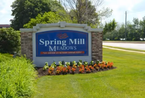 Outdoor view of a large sign for Spring Mill Meadows, an American senior community, surrounded by green grass, colorful flowers, and trees in the background near a road.