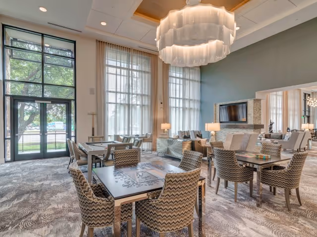 A spacious and well-lit common area with large windows and tall curtains allowing natural light to fill the room. The room features multiple tables with patterned chairs arranged around them, a large modern chandelier hanging from the ceiling, and a seating area with sofas and a wall-mounted television. The decor is contemporary with neutral tones and a patterned carpet.