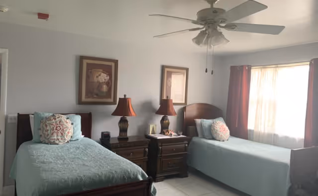 A bedroom with two single beds each covered with light blue bedspreads and decorative pillows. Between the beds are two dark wooden nightstands with matching table lamps. Above the nightstands are two framed floral paintings. A ceiling fan with lights hangs from the ceiling, and sunlight filters through a window with sheer curtains and dark red drapes.