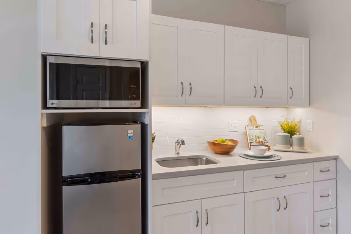 A modern kitchen area with white cabinets, a stainless steel microwave and mini refrigerator, a small sink, and a countertop decorated with a wooden bowl of fruit, a cookbook on a stand, a cup and saucer, and two small potted plants.