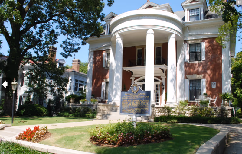 Front exterior view of a large historic brick building with white columns and a curved portico entrance. There is a blue historical marker sign in front of the building, surrounded by a landscaped garden with flowers and greenery. Trees and other buildings are visible in the background under a clear blue sky.
